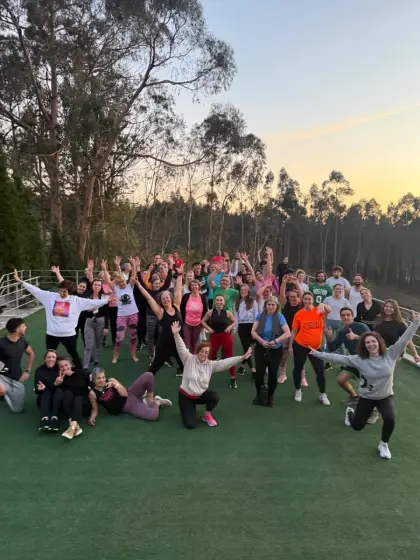 Um grupo grande de alunos no relvado exterior ao pôr do sol, de braços no ar, com os eucaliptos atrás.